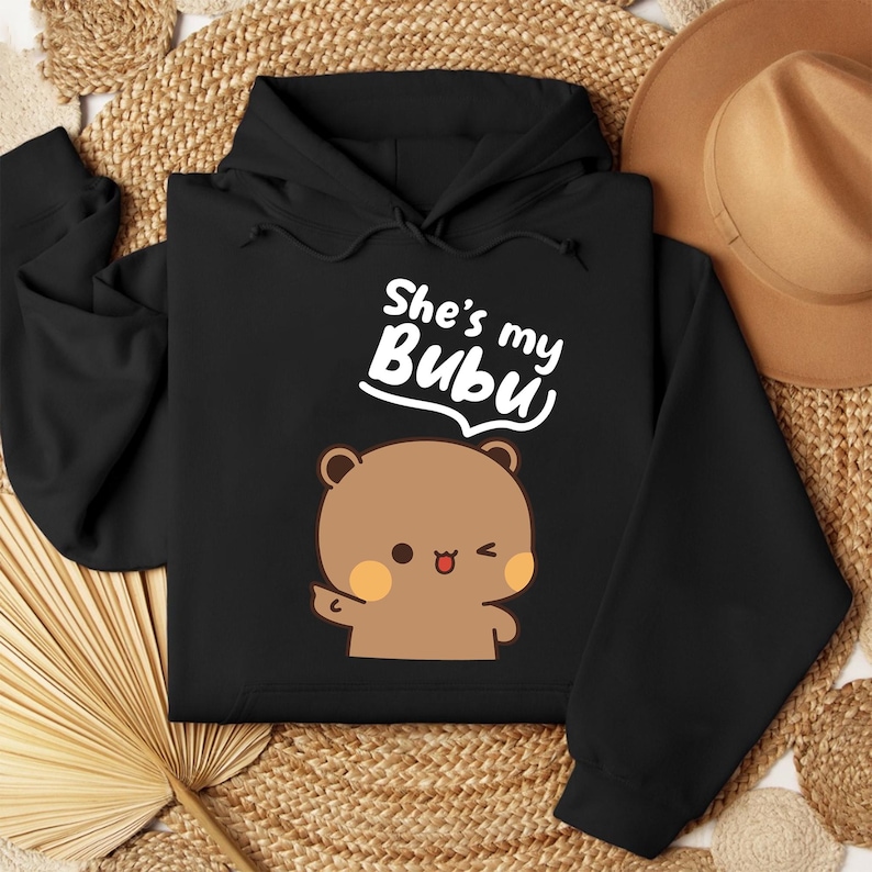 May include: Black hoodie featuring a cartoon bear with a wink and the text "She's my Bubu" in white. The bear is brown with peach-colored cheeks. The hoodie has a drawstring hood and long sleeves.