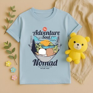 Peut inclure: T-shirt bleu clair avec un motif repr&eacute;sentant des ours dans un hamac et le texte "The Adventure Soul Nomad". Un ours en peluche jaune est plac&eacute; sur le c&ocirc;t&eacute; droit du t-shirt.