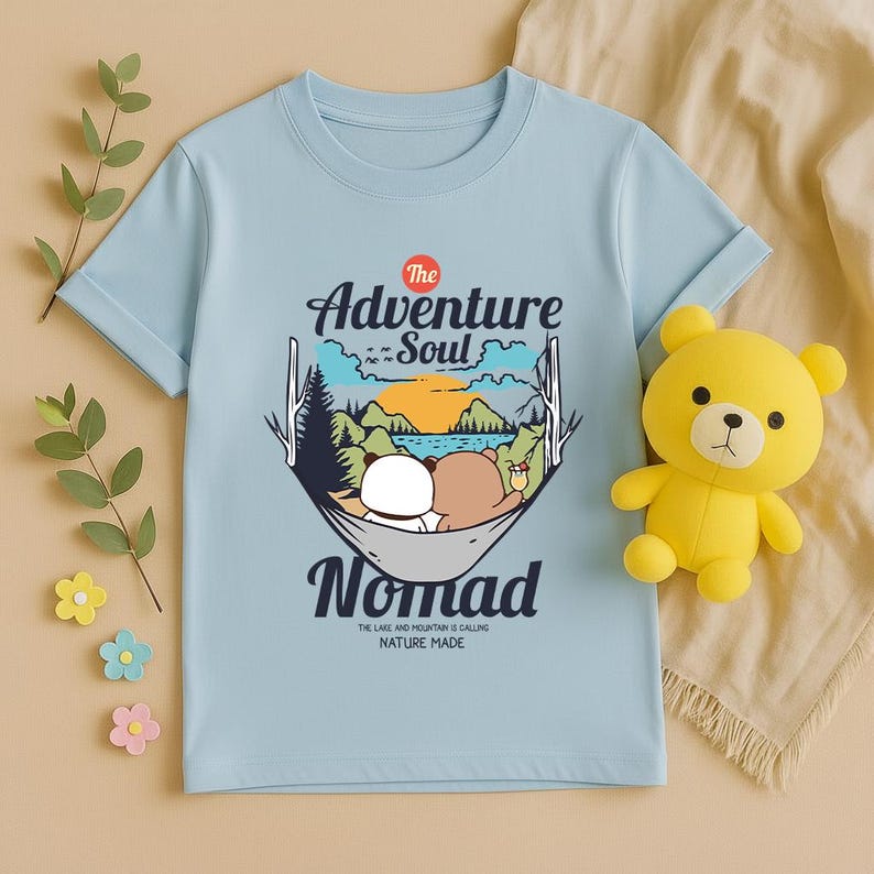 Peut inclure: T-shirt bleu clair avec un motif repr&eacute;sentant des ours dans un hamac et le texte "The Adventure Soul Nomad". Un ours en peluche jaune est plac&eacute; sur le c&ocirc;t&eacute; droit du t-shirt.