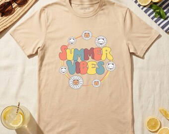 May include: Beige t-shirt with the text "SUMMER VIBES" in colorful bubble letters, surrounded by a daisy chain. The shirt is laid flat on a light-colored surface, with sunglasses and a straw hat in the background.