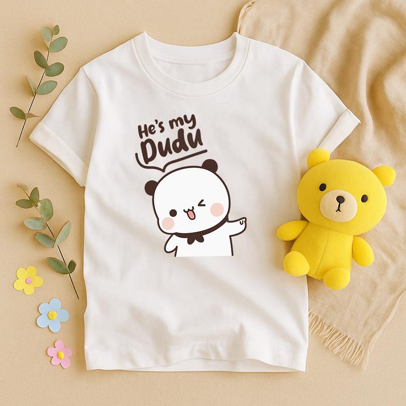 May include: White t-shirt with a cartoon panda illustration and the text "He's my Dudu". A yellow teddy bear is next to the shirt. The shirt has short sleeves and a round neckline. The panda is winking and pointing.