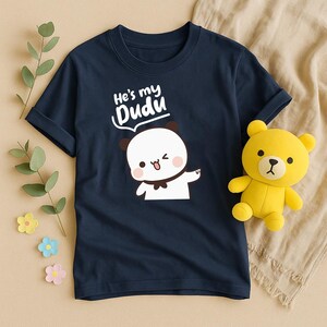 May include: Navy blue t-shirt with the text "He's my Dudu" and a cartoon panda bear graphic. A yellow teddy bear is next to the shirt. The shirt is made of soft material.