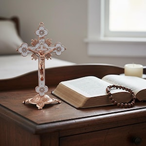 May include: A white and rose gold crucifix with a figure of Jesus Christ, standing on a decorative base. The cross is on a wooden table next to an open book, a rosary, and a lit candle.