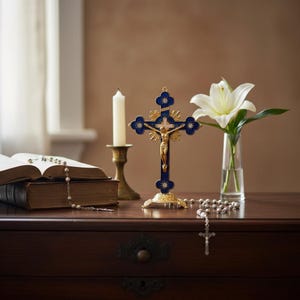 Peut inclure: Une croix bleue et dorée avec une figure de Jésus dorée est posée sur une surface en bois, à côté d'une bougie blanche dans un bougeoir en laiton, d'un livre ouvert et d'un chapelet. Un lis blanc dans un vase en verre est également présent.