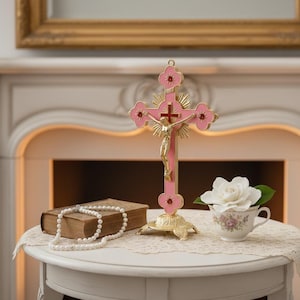 Peut inclure: Une croix rose et dorée avec une figure de Jésus dorée, posée sur une petite table. Un livre ancien, un collier de perles et une tasse à thé avec une rose blanche sont également sur la table. La croix a des accents rouges.