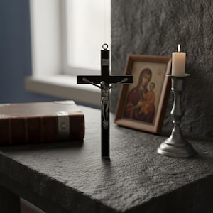 May include: A black crucifix with a silver Jesus figure hangs in front of a window. A lit candle in a silver candlestick, a framed religious icon, and a leather-bound book are also on the dark stone surface.