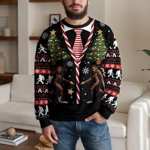 Peut inclure: Un sweat-shirt noir à manches longues avec un motif imprimé représentant un col blanc et une cravate à rayures rouges et blanches. Le design inclut deux silhouettes de Bigfoot, des sapins de Noël, des étoiles dorées et des flocons.