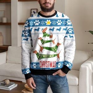 May include: A white and blue long-sleeve sweatshirt featuring a repeating pattern of dachshunds, paw prints, and snowflakes. The centre displays three dachshunds wearing green sweaters, with the text Merry Merry Christmas printed in red.