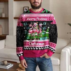 Può includere: Una felpa nera a maniche lunghe con una stampa festiva raffigurante tre fenicotteri rosa con cappelli da Babbo Natale avvolti in luci natalizie. Il testo recita Merry Flocking Christmas e Fla La La La La Mingo, con motivi rosa e bianchi.