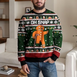 Puede incluir: Una sudadera de cuello redondo verde oscuro con un gráfico de hombre de jengibre con un bastón de caramelo roto y el texto OH SNAP. El diseño incluye un patrón repetitivo de casas de jengibre, árboles de Navidad y copos de nieve blancos.