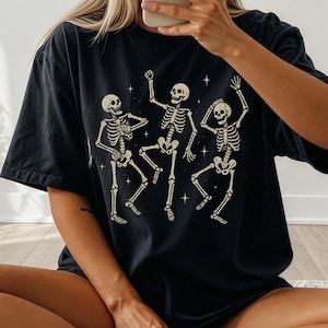 Puede incluir: Camiseta negra con un gráfico beige de tres esqueletos bailando. Los esqueletos están representados en poses dinámicas, con los brazos y las piernas levantadas. Pequeñas estrellas están dispersas alrededor de los esqueletos.