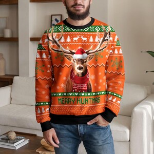 Puede incluir: Una sudadera naranja de manga larga con un gráfico central de un ciervo con un gorro de Papá Noel rojo y una bufanda roja. El diseño incluye copos de nieve blancos, patrones geométricos verdes y blancos, y el texto MERRY HUNTMAS en verde.