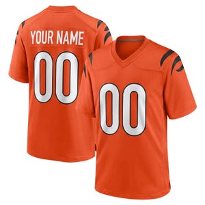 Op de afbeelding: Een oranje shirt met korte mouwen en een V-hals. De voorkant is voorzien van het nummer 00 in het wit met zwarte omlijning. De achterkant toont de tekst YOUR NAME in het wit boven het nummer 00 in het wit met zwarte omlijning en zwarte strepen.