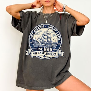 以下が含まれることがあります： チャコールグレーの半袖Tシャツ。帆船が描かれた円形のグラフィックと「LAKE ONTARIO GREAT LAKES」「EST. 1615」「NOT LAKE AMERICA」の文字、そしてバナーの下部には小さなカエデの葉のアイコンがデザインされています。