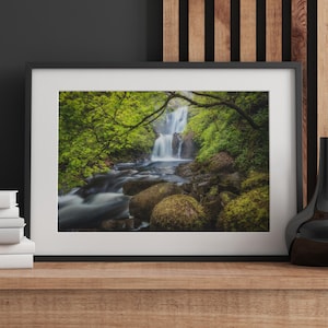 May include: Framed landscape photograph of a waterfall cascading into a river. The image features lush green foliage, moss-covered rocks, and a black frame. The water appears blurred, creating a sense of motion and tranquility.