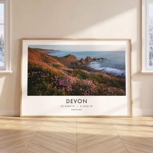 May include: A framed landscape print featuring a coastal scene in Devon, England, with pink wildflowers on a cliffside overlooking the ocean. The text below the image reads: DEVON, 50.9940°N | 4.5326°W, ENGLAND, in a clean, black sans-serif font.