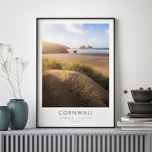 May include: A framed print depicting a coastal scene in Cornwall, England. The photograph shows a sandy beach, grassy dunes, and the sea under a bright sky. The word "Cornwall" is printed in white at the bottom, with coordinates.
