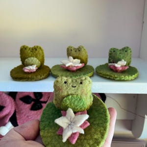 May include: Handmade felt frog figurines. Each frog is light green with a smiling face, sitting on a green lily pad with a white and pink flower. Three frogs are on a white shelf, and one is held in a hand.
