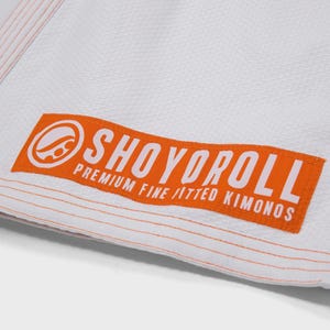 Op de afbeelding: Close-up van een wit vechtsportuniform met oranje stiksels. Een oranje rechthoekige patch met de merknaam "SHOYOROLL" in witte letters is op het uniform genaaid. De patch bevat ook de tekst "PREMIUM FINE FITTED KIMONOS".