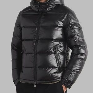 Puede incluir: Una chaqueta acolchada negra brillante con capucha y cremallera completa. La chaqueta tiene un diseño acolchado, un bolsillo en la manga izquierda y un pequeño bolsillo en el pecho. Diseñada para el clima frío.