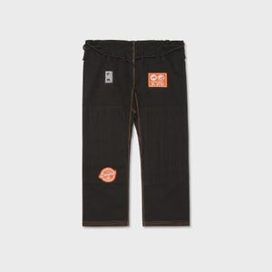 Op de afbeelding: Zwarte vechtsportbroek met oranje en witte patches. De broek heeft bruine stiksels en een tailleband met trekkoord. Patches bevatten tekst en cirkelvormige ontwerpen. De broek is gemaakt van een duurzaam materiaal.