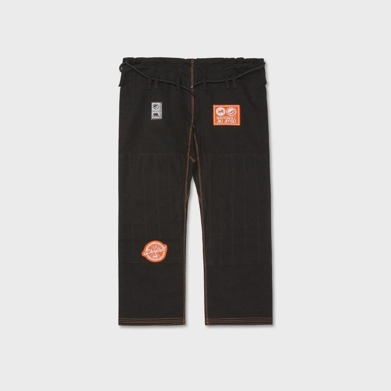 Op de afbeelding: Zwarte vechtsportbroek met oranje en witte patches. De broek heeft bruine stiksels en een tailleband met trekkoord. Patches bevatten tekst en cirkelvormige ontwerpen. De broek is gemaakt van een duurzaam materiaal.