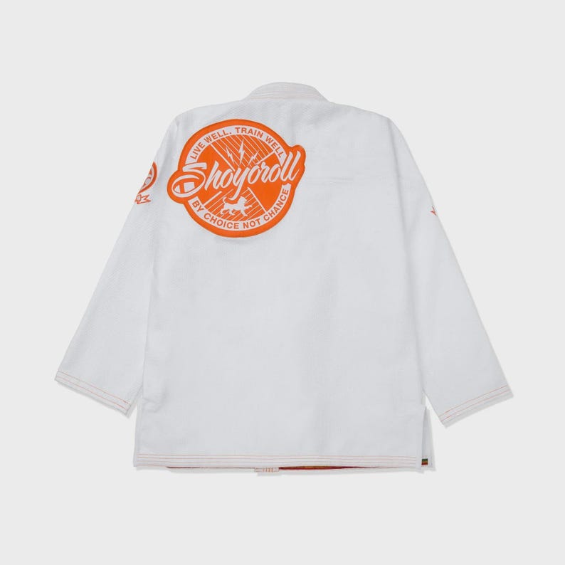 Op de afbeelding: Witte vechtsportuniform met oranje accenten. De achterkant heeft een groot oranje cirkelvormig logo met de tekst "Shoyoroll" en de zinnen "Live Well, Train Well" en "By Choice Not Chance". Het uniform heeft oranje stiksels.