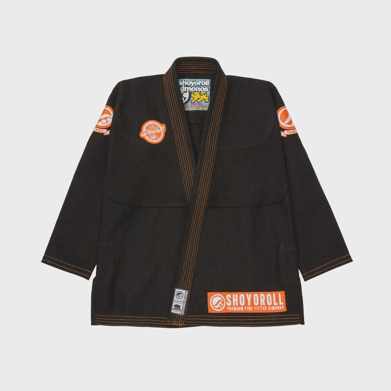Op de afbeelding: Zwarte Jiu-Jitsu gi met oranje stiksels en patches. De gi heeft het "SHOYOROLL" logo op de borst, schouders en rok. De gi is gemaakt van een duurzame stof en is ontworpen voor vechtsporttraining en -competitie.