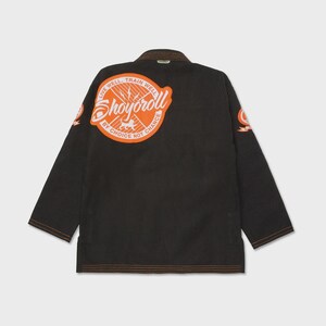 Op de afbeelding: Zwarte Jiu-Jitsu gi met oranje en witte logo patches. De achterkant heeft een grote ronde patch met de tekst "Shoyoroll" en de zin "Live Well, Train Well, By Choice Not Chance". De gi heeft oranje stiksels.