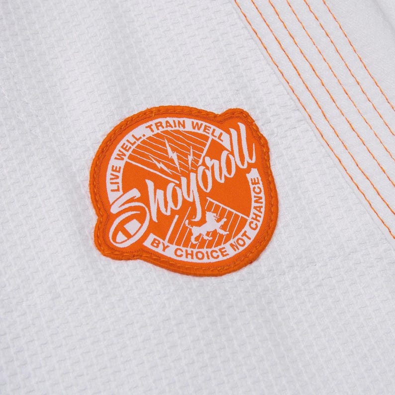 Op de afbeelding: Close-up van een wit vechtsportuniform met een oranje patch. De patch bevat het woord "Shoyoroll" in het wit, omringd door tekst en een afbeelding. Oranje stiksels accentueren het uniform.