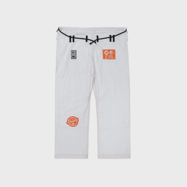 Op de afbeelding: Witte vechtsportbroek met zwarte details in de taille en oranje patches. De broek heeft een trekkoordsluiting en geborduurde patches met logo's en tekst. Ontworpen voor vechtsporttraining en -wedstrijden.