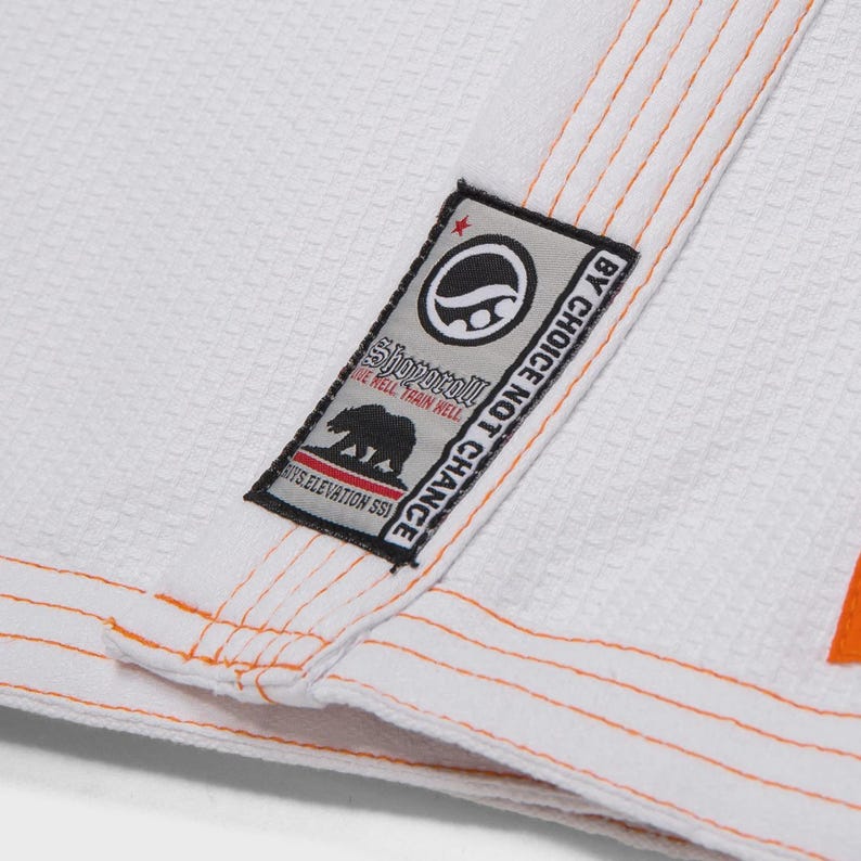 Op de afbeelding: Close-up van een wit vechtsportuniform met oranje stiksels en afwerking. Een rechthoekige patch toont een logo en de tekst "BY CHOICE NOT CHANCE". Het uniform heeft een geweven textuur.