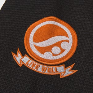 Op de afbeelding: Een oranje en witte geborduurde patch met de tekst "LIVE WELL" op een banner. De patch heeft een cirkelvormig ontwerp met een golf en twee cirkels. De patch zit op een donkergrijze, getextureerde stof.
