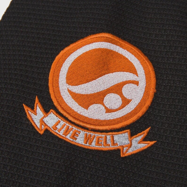 Op de afbeelding: Een oranje en witte geborduurde patch met de tekst "LIVE WELL" op een banner. De patch heeft een cirkelvormig ontwerp met een golf en twee cirkels. De patch zit op een donkergrijze, getextureerde stof.