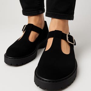 Może przedstawiać: Czarne buty z paskiem w kształcie litery T z grubą podeszwą i zapięciem na klamrę. Buty mają fakturę przypominającą zamsz i okrągły nosek. Podeszwa jest gruba i ma bieżnik. Noszone z czarnymi spodniami.