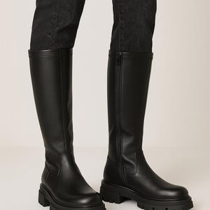 Könnte beinhalten: Schwarze kniehohe Stiefel mit einer klobigen Profilsohle. Die Stiefel haben eine glatte Oberfläche und einen seitlichen Reißverschluss. Getragen mit schwarzen Jeans.