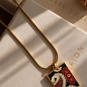 Könnte beinhalten: Eine goldfarbene Kettenhalskette mit einem rechteckigen Anhänger auf schwarzem Emaille-Hintergrund, verziert mit einem roten und cremefarbenen Paisley-Muster. Der Anhänger ist an einer dünnen, flexiblen goldfarbenen Schlangenkette befestigt.