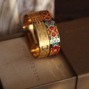 以下が含まれることがあります： NASK COLLECTIONと記されたベージュの箱の上に置かれた2つのゴールドトーンのバングル。片方のバングルには精巧なエンボス加工のフローラルパターンが施され、もう片方には赤、青、白のフローラルモチーフがあしらわれた鮮やかなマルチカラーのエナメルデザインが施されています。