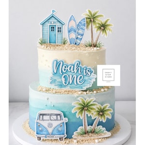 May include: A two-tiered cake with blue ombre frosting decorated with beach-themed paper toppers including a blue beach hut, two surfboards, palm trees, and a vintage van. The text "Noah is one" is displayed on the top tier in blue script.