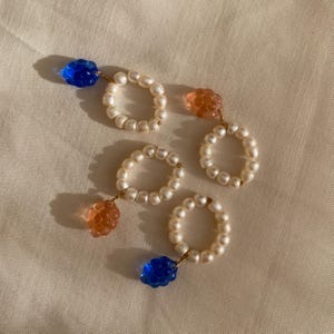 Può includere: Quattro anelli con perle bianche e ciondoli colorati. Due anelli hanno un ciondolo blu a forma di fiore, e due hanno un ciondolo arancione a forma di bacca. Gli anelli sono su una superficie color crema.