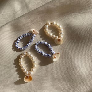 May include: Four handmade rings. Two rings feature white pearls and small seashells. The other two rings have blue and white striped beads with small seashells. The rings are on a cream-colored fabric.