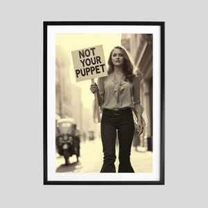 Puede incluir: Impresión enmarcada en tonos sepia que muestra a una persona sosteniendo un cartel con el texto «NOT YOUR PUPPET». La persona lleva una blusa, pantalones oscuros y un cinturón. El fondo muestra una escena callejera con un vehículo.