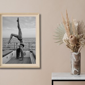 Peut inclure: Photographie encadr&eacute;e en noir et blanc d'une personne faisant le poirier sur le pont d'un bateau, avec l'oc&eacute;an et le ciel en arri&egrave;re-plan. La personne porte un bikini noir. L'image est dans un cadre en bois clair.
