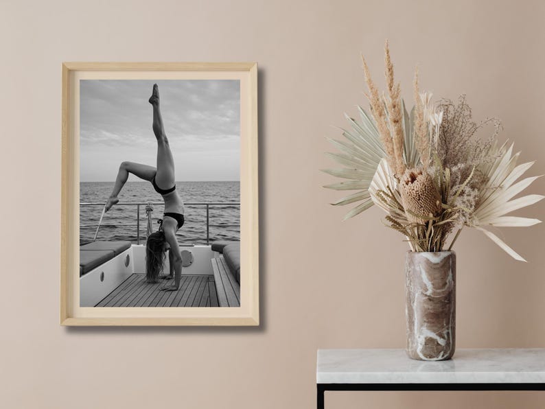 Peut inclure: Photographie encadr&eacute;e en noir et blanc d'une personne faisant le poirier sur le pont d'un bateau, avec l'oc&eacute;an et le ciel en arri&egrave;re-plan. La personne porte un bikini noir. L'image est dans un cadre en bois clair.
