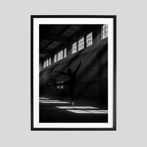 May include: Black and white framed print of a ballerina in a large, industrial space. The dancer is in mid-leap, arms outstretched, with sunlight streaming through the windows. The image is a study in contrasts, with the dancer's silhouette against the bright light.
