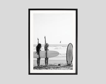 May include: Black and white framed print of two people standing on a beach, holding a surfboard. They are facing the ocean, with their arms raised in a peace sign. A second surfboard stands upright nearby.