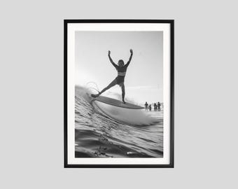 May include: Monochrome framed print showing a surfer on a wave. The surfer is a silhouette with arms raised. Other figures are visible in the distance. The image captures the dynamic action of surfing, set against a bright sky.