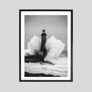 May include: A black and white framed photograph of a tall lighthouse standing on a rocky shore. Large, crashing ocean waves spray white water around the base of the structure. The sky is overcast and grey, creating a high-contrast scene.