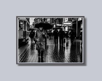 May include: Black and white photograph of a rainy city street scene. A person in a long coat and holding a black umbrella walks toward the viewer. Other people with umbrellas are in the background, along with a tram and storefronts.