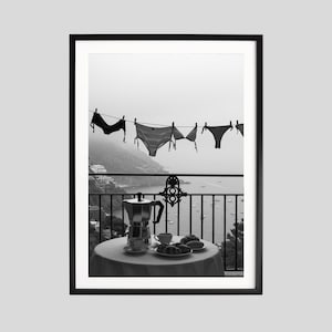 Op de afbeelding: Zwart-witfoto van een balkonsc&egrave;ne. Ondergoed hangt aan een waslijn met uitzicht op een kustlandschap. Een tafel is gedekt met koffie, gebak en een koffiezetapparaat. De afbeelding is ingelijst.